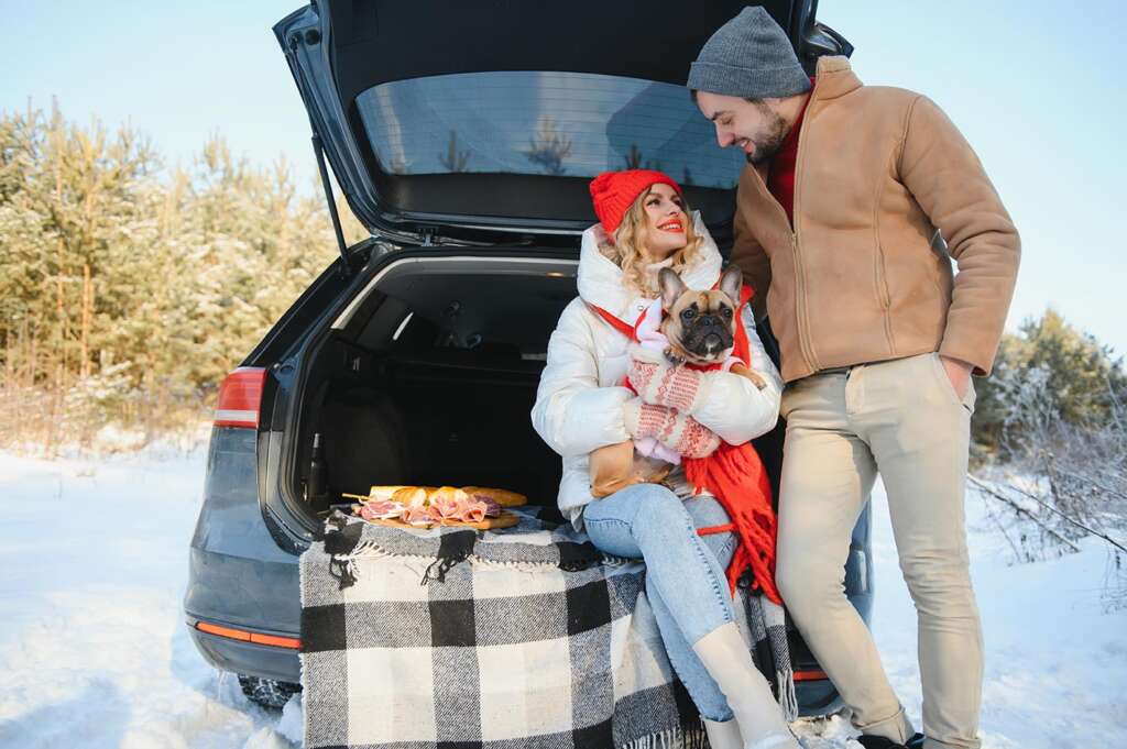 A happy couple and their dog sitting in the open back of a car having a winter picnic on a wood charcuterie board